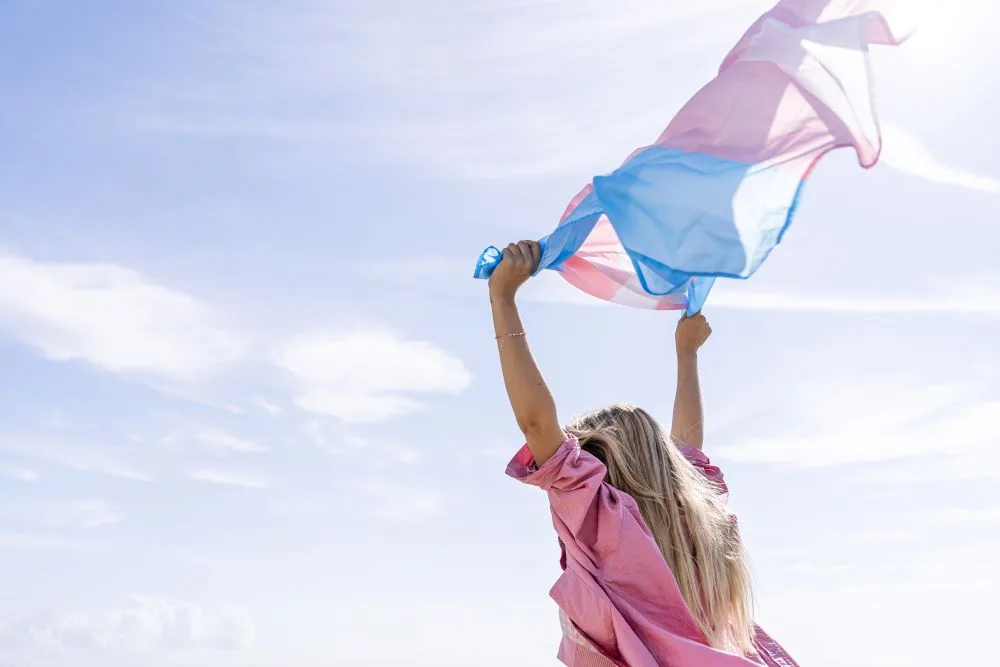 A transwoman holding a transgender flag in the air with joy.