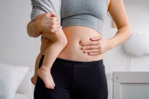 A woman holding her child in one arm and touching her abdomen where her C-Section scar is with the other.
