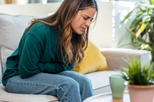 Young woman leaning on the couch in the living room at home while holding her stomach in pain.